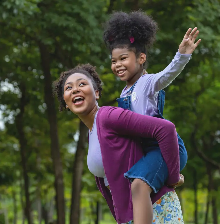 Mom laughing as she carries her young daughter piggyback through the park.